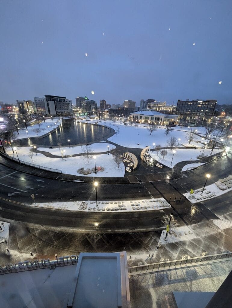 Snowy early morning view overlooking Big Spring International Park in Downtown Huntsville, Alabama, as seen from the accessible room at the AC Hotel. The park, roads, and nearby buildings are gently lit and blanketed in snow, creating a peaceful winter cityscape.