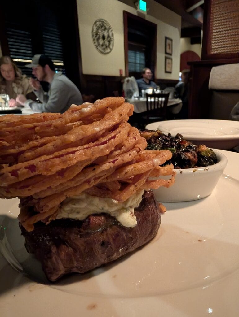 A perfectly cooked filet mignon topped with creamy Boursin cheese and a towering stack of crispy tobacco onions, served with a side of roasted Brussels sprouts at Connors Steak & Seafood in Huntsville, Alabama.