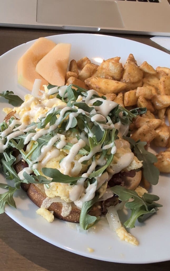 A savory breakfast plate from the AC Hotel Downtown Huntsville featuring scrambled eggs, arugula, and creamy dressing over toast, with a side of seasoned breakfast potatoes and fresh cantaloupe slices.
