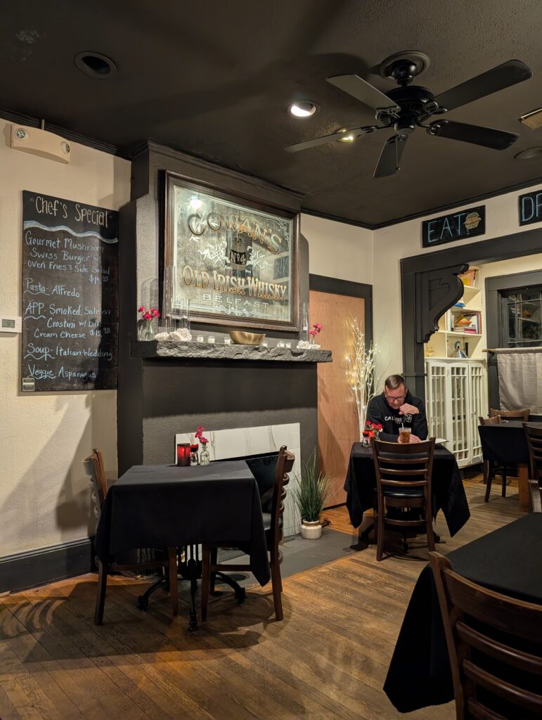 A warm, intimate dining room at Rock House Eatery in Guntersville, Alabama, featuring dark wood floors, black tablecloths, and vintage-style decor including a “Cowan’s Old Irish Whisky” mirror and chalkboard menu of daily specials.