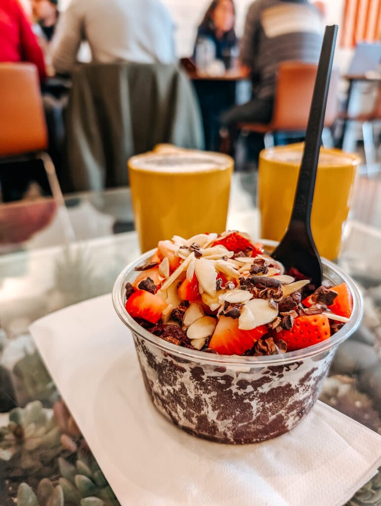 Close-up of an acai bowl topped with sliced strawberries, almonds, and cacao nibs on a glass table at Honest Coffee Roasters in Downtown Huntsville, Alabama. Two yellow mugs of latte sit in the background with a cozy café crowd slightly out of focus.
