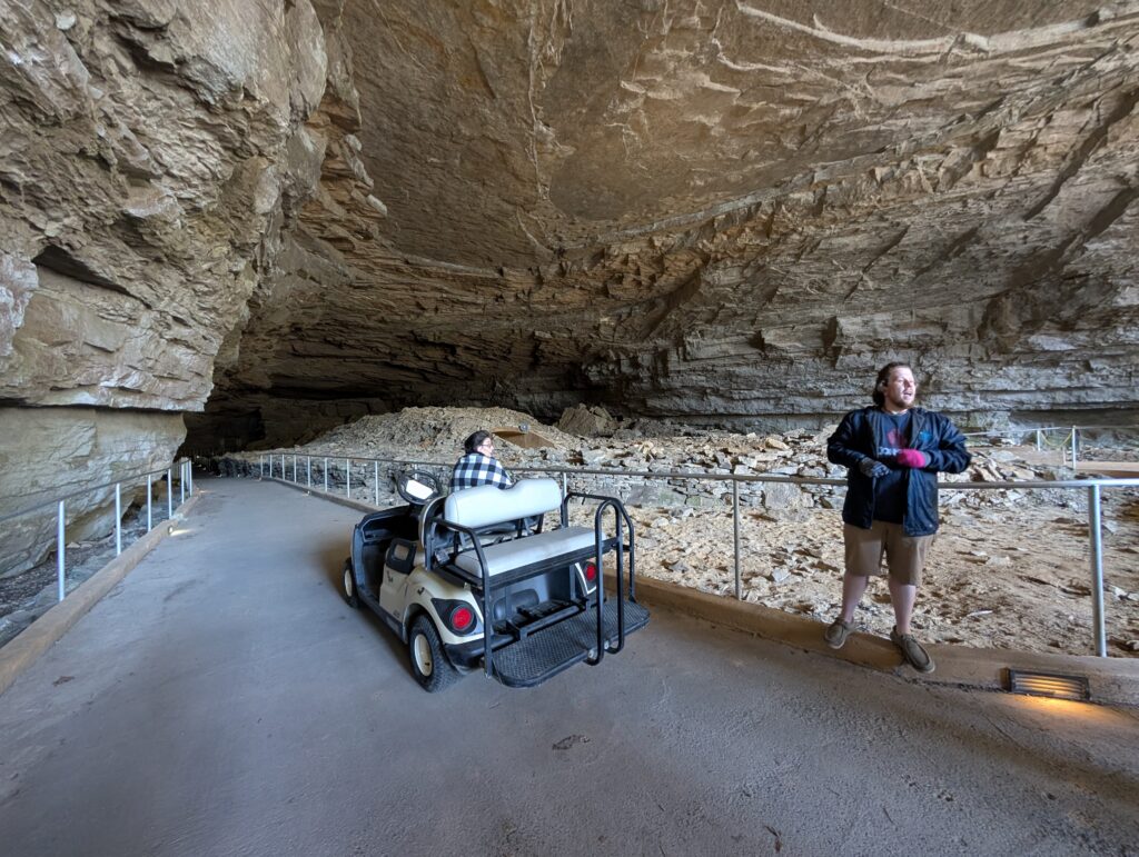 A tour guide stands beside a golf cart at the entrance of Cathedral Caverns in Alabama, surrounded by towering rock formations and a paved, accessible path that winds through the massive cave system.