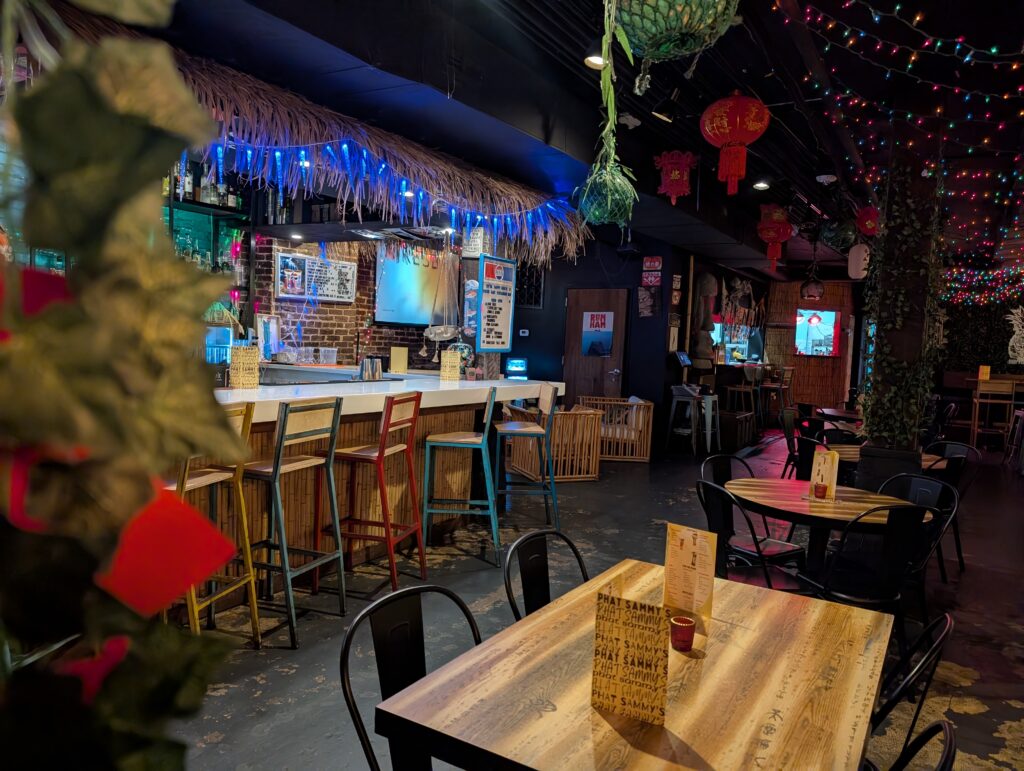Colorful interior of Phat Sammy’s in downtown Huntsville, Alabama, featuring tropical decor, string lights, Asian lanterns, and a mix of bamboo and metal furnishings with vibrant neon signage behind the bar.