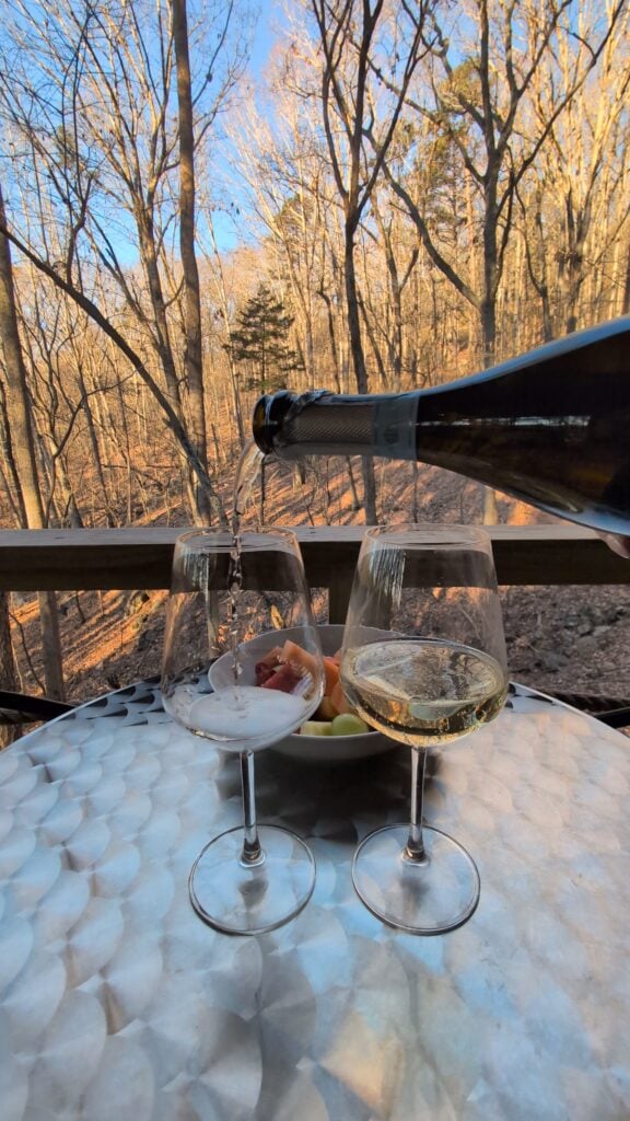A bottle of sparkling wine is poured into one of two glasses set on a round table at ReTreet Resort and Spa in Lake Guntersville, Alabama. A bowl of fresh fruit sits between the glasses, with a backdrop of a peaceful winter forest under a clear blue sky.