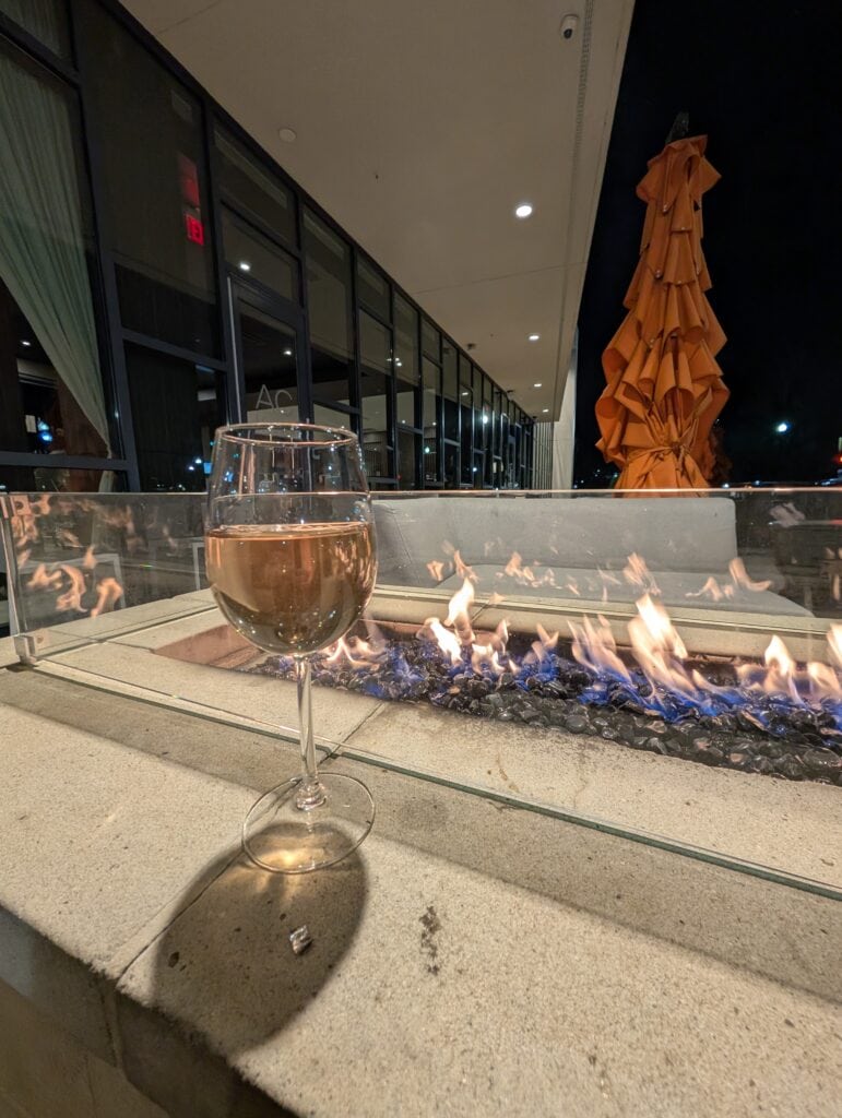 A glass of rosé wine on the edge of a stone fire pit with blue flames glowing, set outside the AC Hotel Downtown Huntsville at night with an orange umbrella in the background.