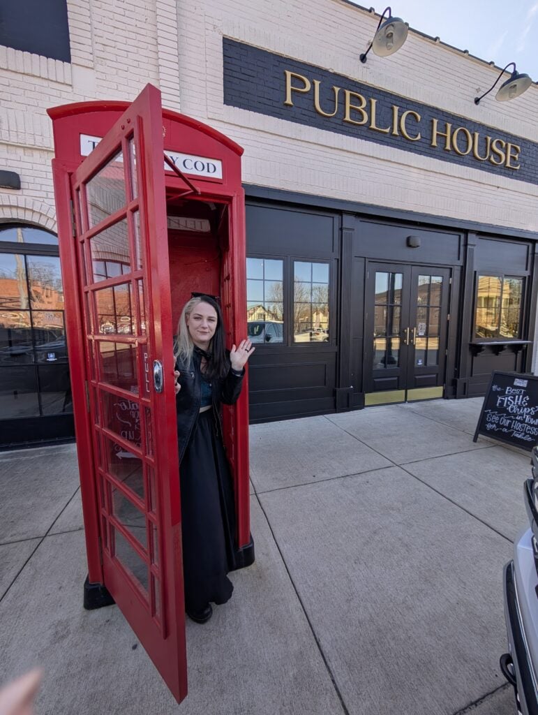 Phoenyx stands in a bright red British-style phone booth outside The Salty Cod Public House in Athens, Alabama, smiling and waving. The pub’s sign and a chalkboard advertising fish and chips are visible in the background.