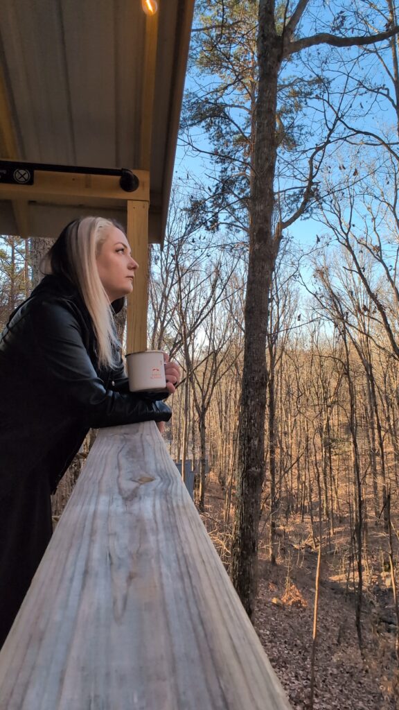 Phoenyx leans on the wooden railing of a glamping tent porch at ReTreet Resort and Spa in Lake Guntersville, Alabama, sipping coffee and gazing thoughtfully into the surrounding winter forest of bare trees under a clear blue sky.