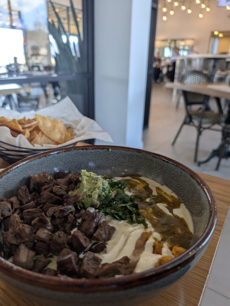A hearty steak bowl at La Esquina Cocina in Guntersville, Alabama, featuring grilled steak, guacamole, fresh cilantro, creamy sauces, and salsa verde, served with crispy tortilla chips in a modern, airy dining space.