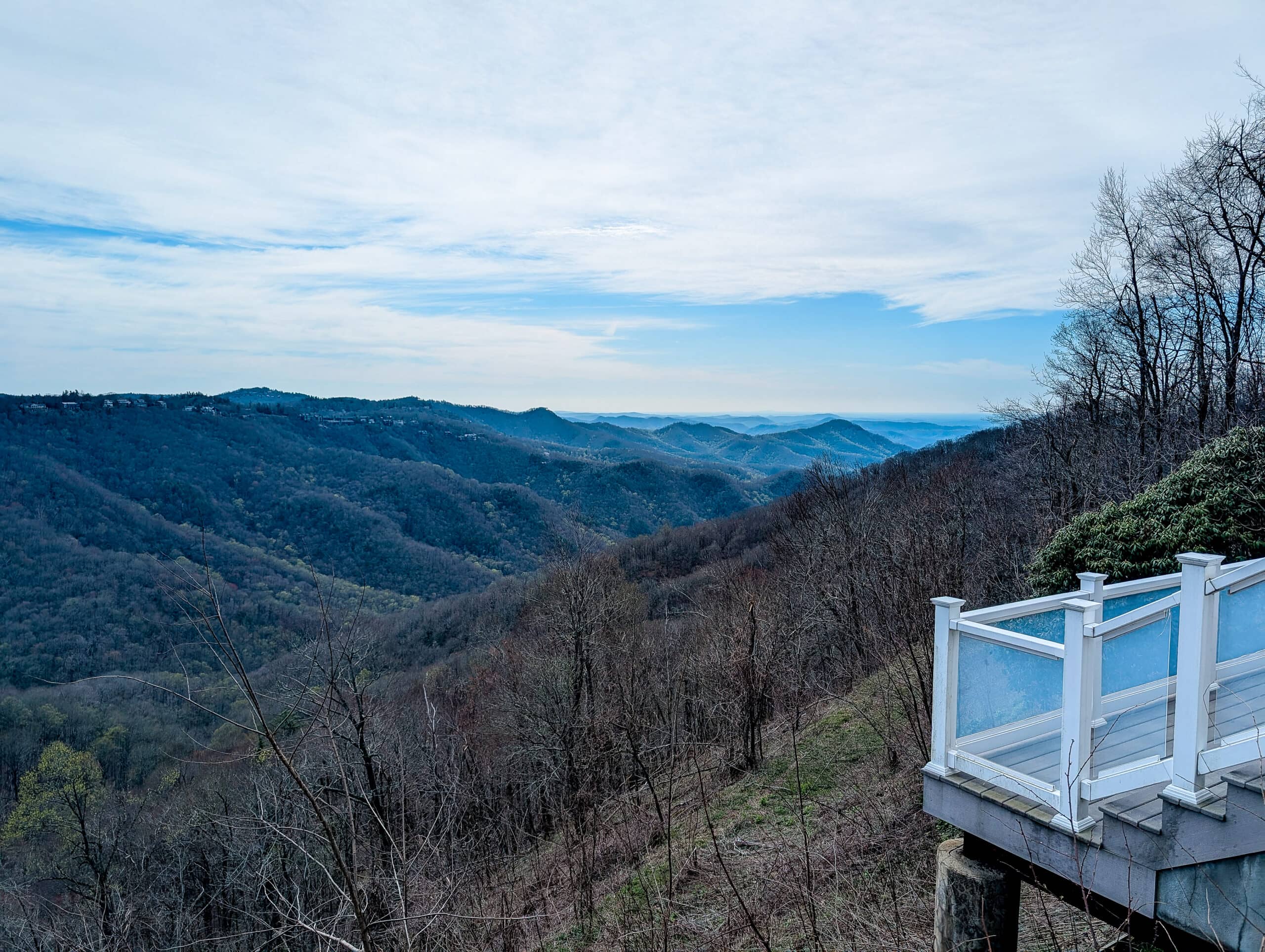 A breathtaking view from Westglow Spa reveals layers of rolling Blue Ridge ridges fading into the horizon. The wooden deck with white railings offers a peaceful vantage point for soaking in the mountain serenity for the perfect weekend in Blowing Rock.