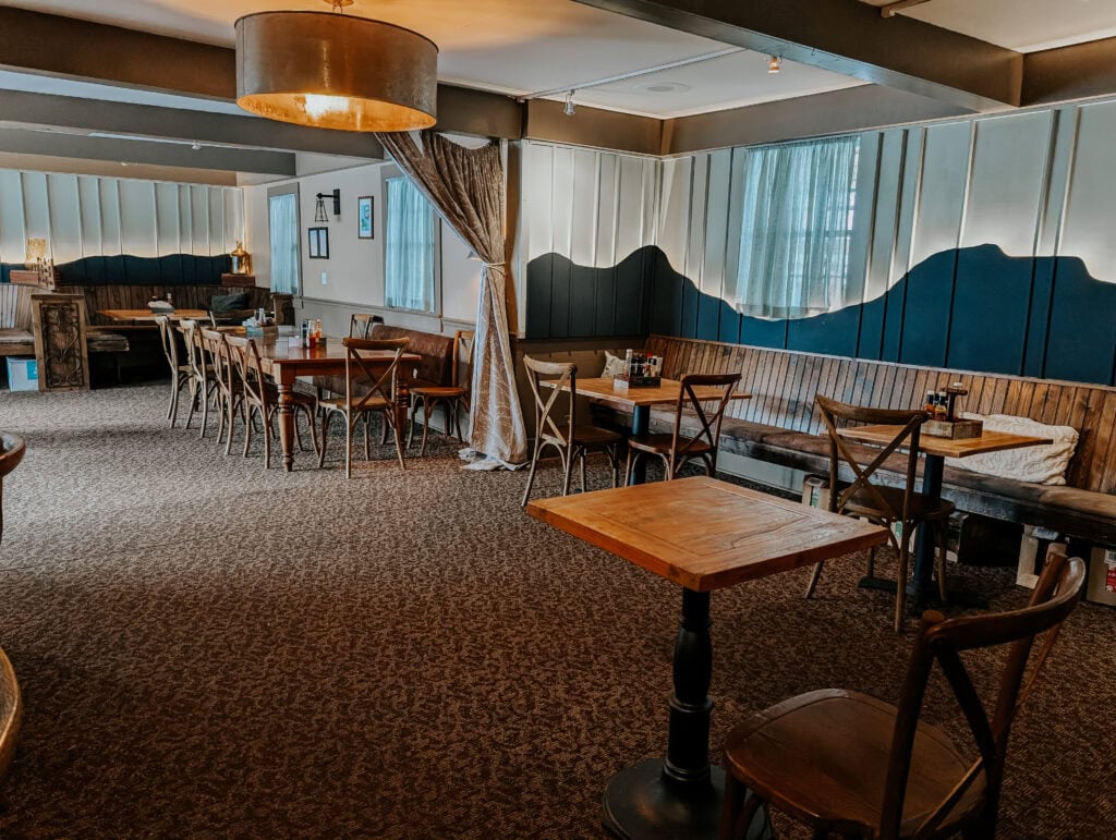 The dining area at Hellbender Bed and Beverage blends rustic mountain style with laid-back comfort. Wood-paneled banquette seating lines the wall beneath a mural-style design reminiscent of rolling Blue Ridge ridges, while wooden tables and warm lighting create an inviting, homey atmosphere perfect for lingering over a hearty Southern-inspired meal.