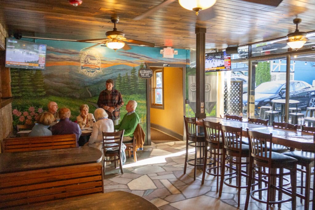 A cozy group of patrons enjoys conversation and craft brews inside the welcoming dining area of Blowing Rock Brewery. Wood-paneled booths and bar stools line the space, while the colorful mural of the Blue Ridge Mountains and the brewery’s logo sets a distinctly local tone. Soft light pours through the large front windows, making the taproom feel bright and laid-back.