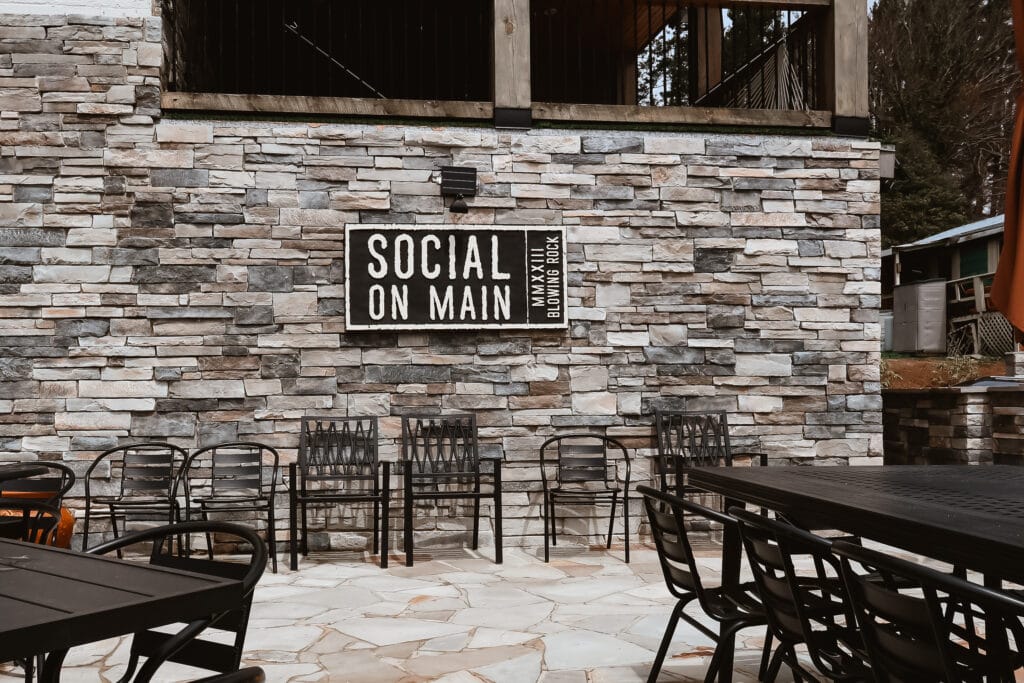 Outdoor seating area at Social on Main restaurant in Blowing Rock, NC, featuring black metal chairs and a stone wall with a bold black and white sign.