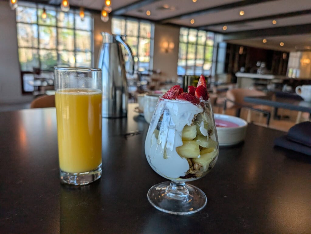 Breakfast scene at The Embers Hotel featuring a glass of orange juice and a parfait with strawberries, pineapple, yogurt, and granola, set on a dark table in a sunlit dining room.