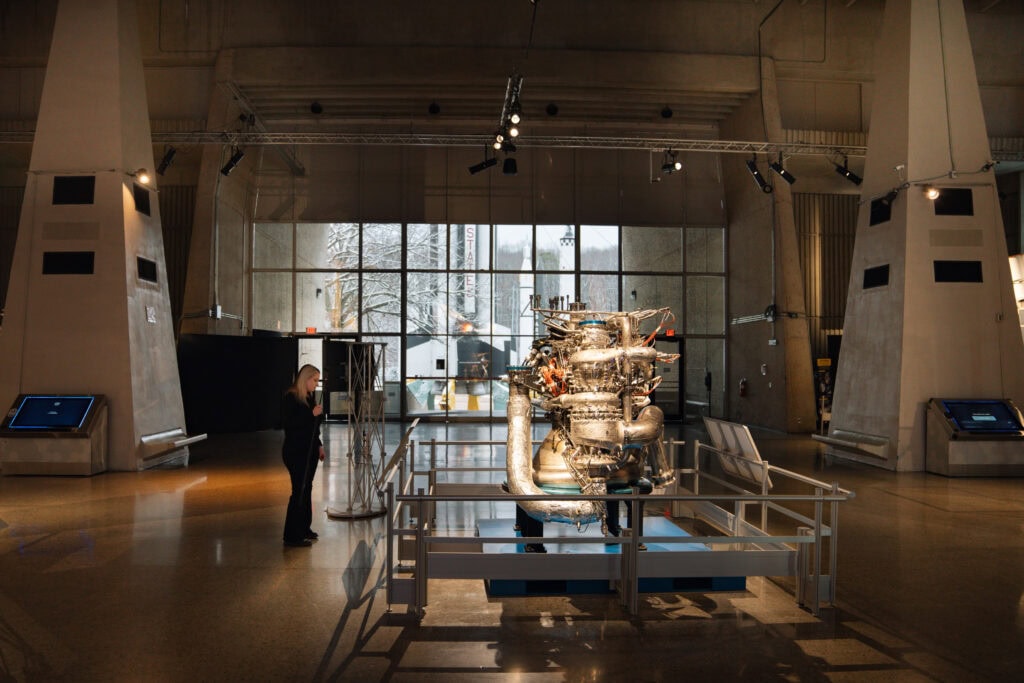 Phoenyx stands in front of a detailed rocket engine exhibit at the Davidson Center for Space Exploration, with snowy outdoor scenes visible through large glass windows. This photo, courtesy of AMLA via The Library Of, captures the high-ceilinged, industrial-style interior of the museum, with additional interactive displays nearby.
