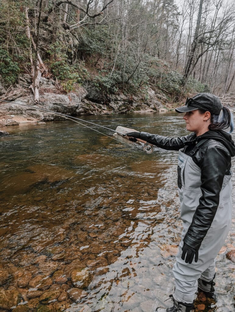 Phoenyx dressed in waders and a cap casts her line into a calm mountain stream while fly fishing with Chetola.
