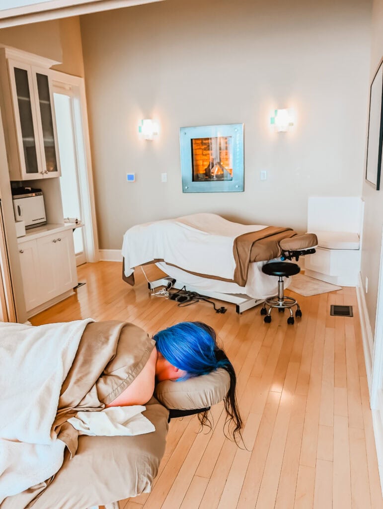Phoenyx relaxes face down on a massage table wrapped in cozy linens, ready to unwind in a tranquil room warmed by a wall-mounted fireplace. The peaceful scene captures a moment of restorative care.