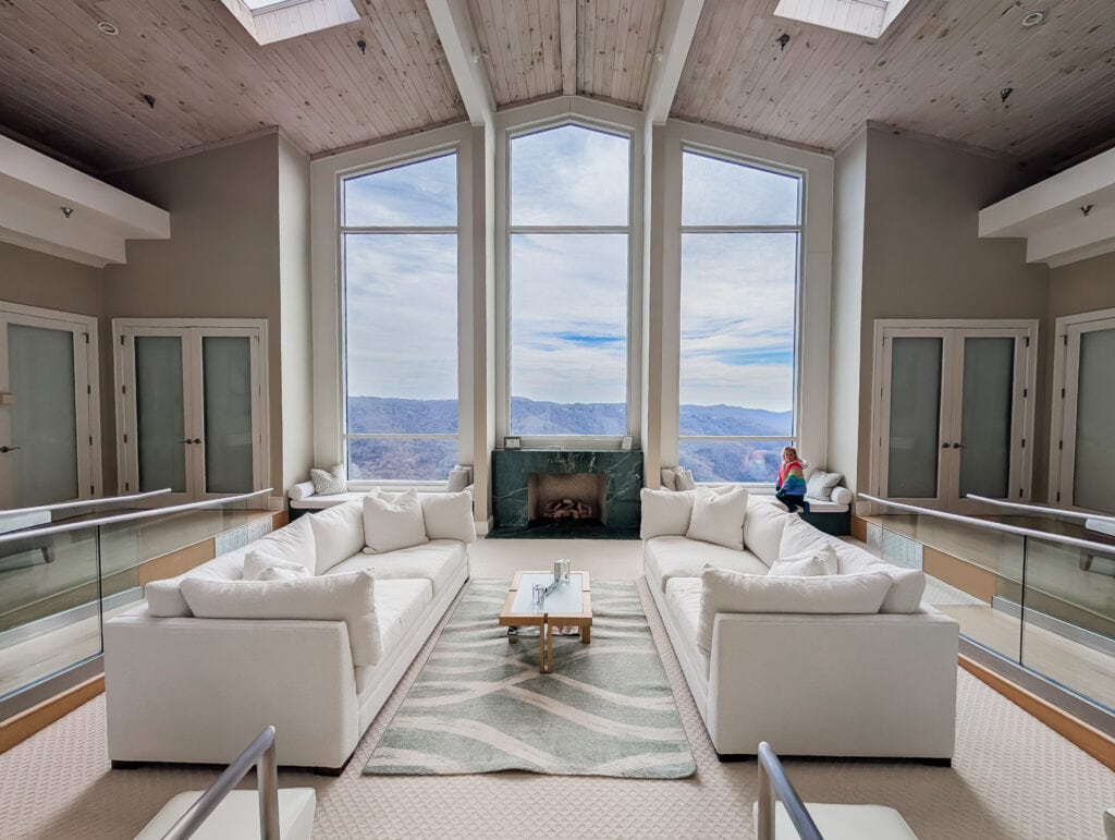 The tranquil lounge at Westglow Spa features oversized white sofas, a glowing fireplace, and soaring windows framing panoramic mountain views. A quiet moment here is the perfect pause between treatments.