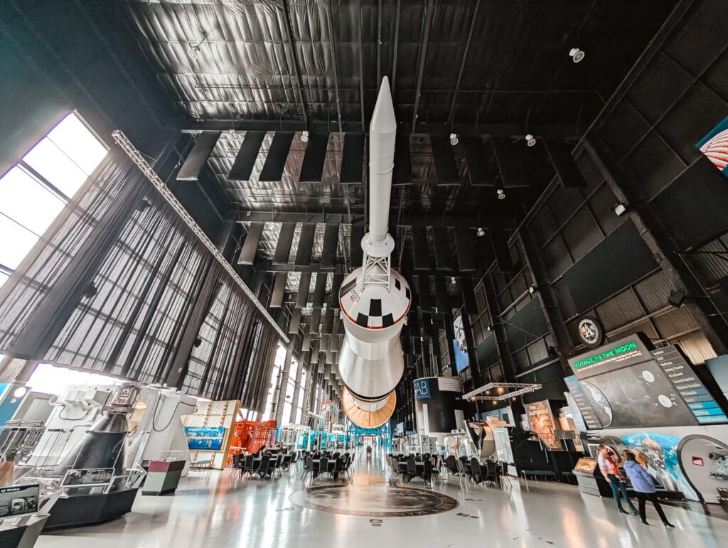 The massive Saturn V rocket suspended overhead in the spacious Rocket Park at the US Space and Rocket Center, surrounded by exhibit displays and educational installations about space exploration and lunar missions.