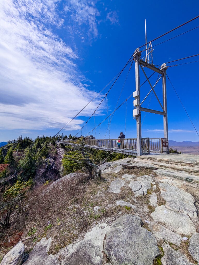 Grandfather Mountain’s iconic suspension bridge stretches across rocky cliffs under a bright blue sky. The area is partially accessible but includes uneven terrain leading up to the bridge.