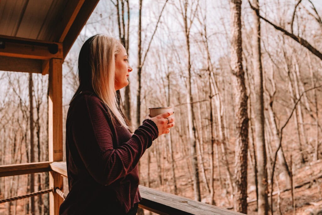 Woman holding a mug stands on a wooden cabin porch, gazing out at a quiet, leafless forest in early winter at ReTreet Resort + Spa in Lake Guntersville, Alabama. Featured image for the blog post Overcoming Mom Guilt: Why Solo Travel is Self-Care.