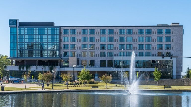 AC Hotel by Marriott in downtown Huntsville sits beside a pond with a water fountain and landscaped park area on a clear day. The modern glass and brick building overlooks walking paths and green space in the city center.