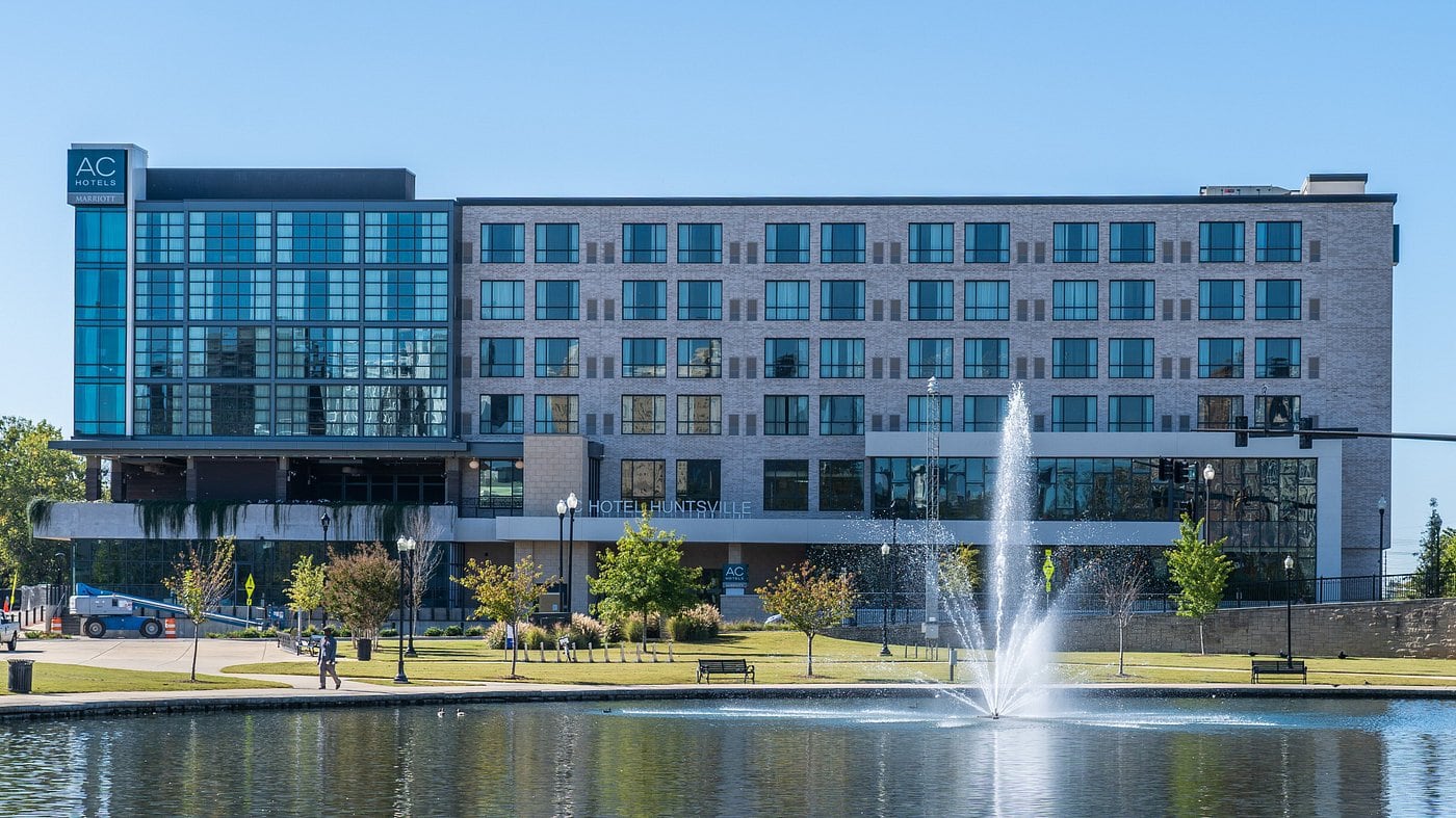 AC Hotel by Marriott in downtown Huntsville sits beside a pond with a water fountain and landscaped park area on a clear day. The modern glass and brick building overlooks walking paths and green space in the city center.