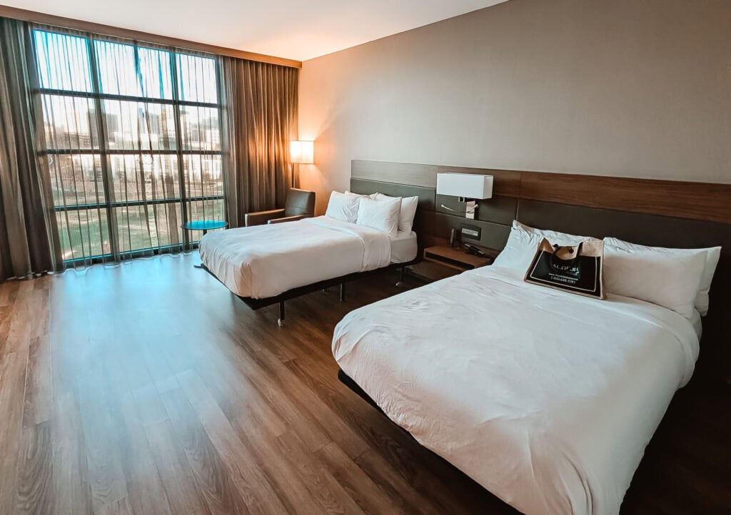 Accessible hotel room at the AC Hotel in downtown Huntsville, featuring two double beds with crisp white linens, hardwood floors for easy wheelchair mobility, wide open space, bedside outlets, and a floor-to-ceiling window offering natural light and city views.