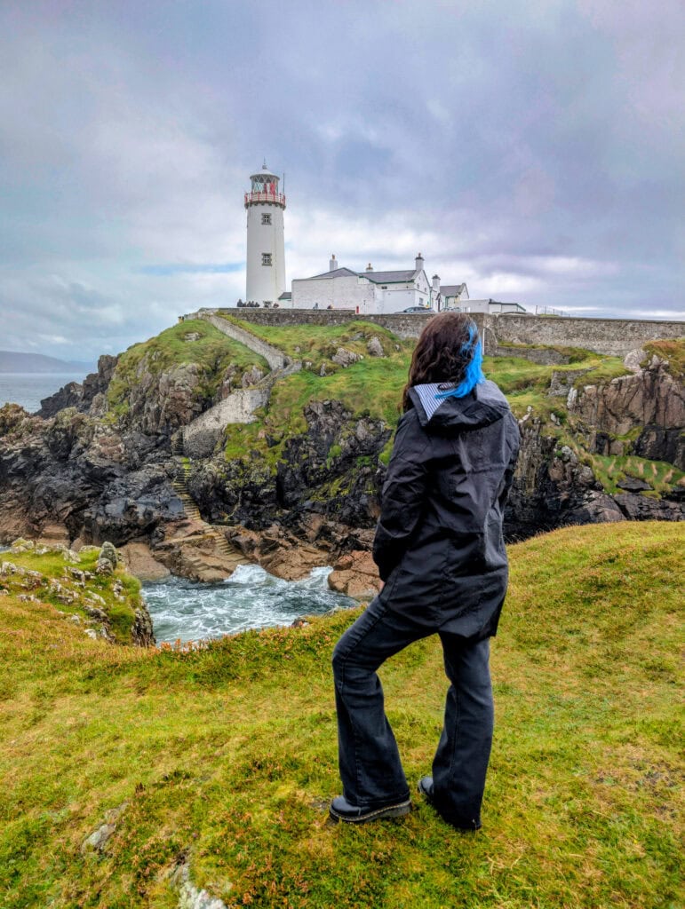 A woman in a black jacket stands on a grassy cliffside overlooking the dramatic coastline and whitewashed Fanad Head Lighthouse in Ireland. Waves crash against the rocks below under a cloudy sky, creating a moody, windswept atmosphere. Featured image for About Page on Phoenyx Travels