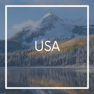Square graphic labeled “USA” with a scenic background of a snowy mountain, pine trees, and a reflective lake. Hints of yellow autumn foliage peek through the evergreens under a crisp blue sky.