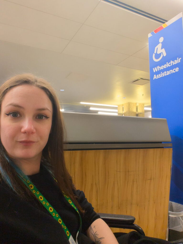 Woman sitting in an airport wheelchair near a counter with a blue sign that reads “Wheelchair Assistance” while waiting for Southwest airport assistance.