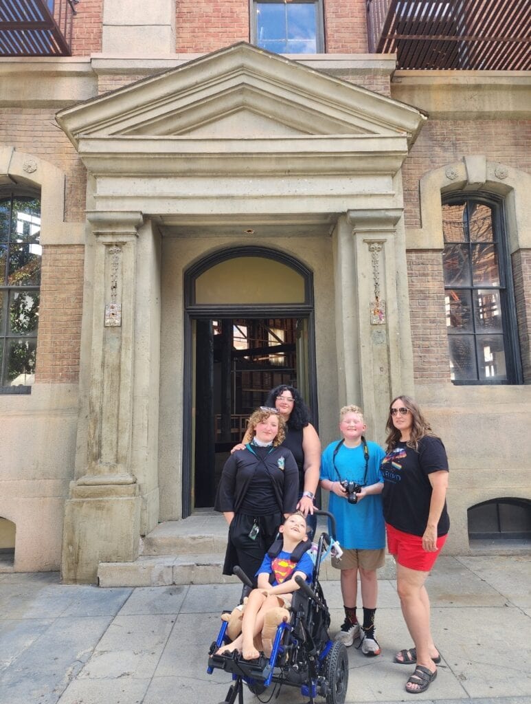 Amy Tarpein with her children and son Elijah in a wheelchair outside the orphanage from the movie, Annie, representing accessible family travel and the wheelchair van fundraiser