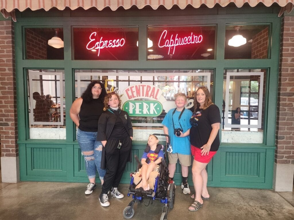 Amy Tarpein and her family with Elijah in his wheelchair in front of Central Perk, highlighting accessible family travel and the Flight Ready Travel Kit fundraiser