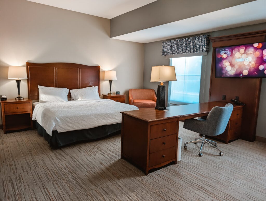 Accessible King Suite at Hampton Inn & Suites Jacksonville NC featuring a king sized bed with bedside tables on each side with a lamp, a desk, and a chair.