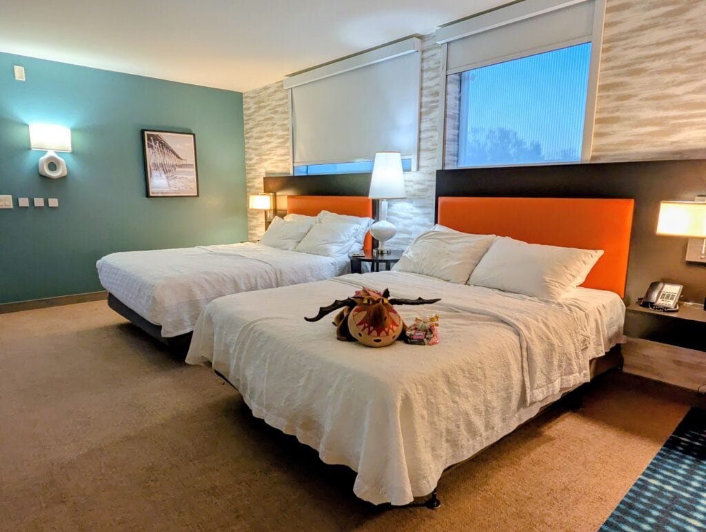 A bright accessible hotel room at Home2Suites Jacksonville that has two large beds with crisp white bedding and vivid orange upholstered headboards beneath tall windows. A playful stuffed dragon and a small bag of treats rest on the nearest bed while warm lamps and a teal accent wall make the room feel colorful and welcoming.
