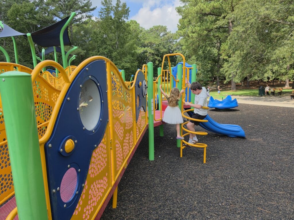A young girl in a white dress and an older boy climb bright yellow spiral bars on a colorful playground surrounded by tall green trees at Northeast Creek Park in Jacksonville, NC. The play structure is filled with curved orange railings green posts blue panels and a blue slide over a soft black ground surface.