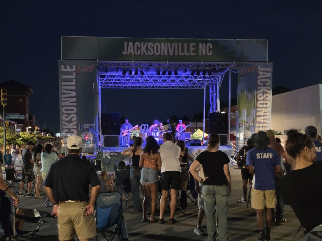 A live band performs beneath blue stage lights at the Rock the Block event in Downtown Jacksonville NC while a crowd gathers in the street to watch. The stage is framed by large signs reading "JACKSONVILLE NC" and "JACKSONVILLE Live" with Jacksonville Recreation and Parks branding visible around the performance area.