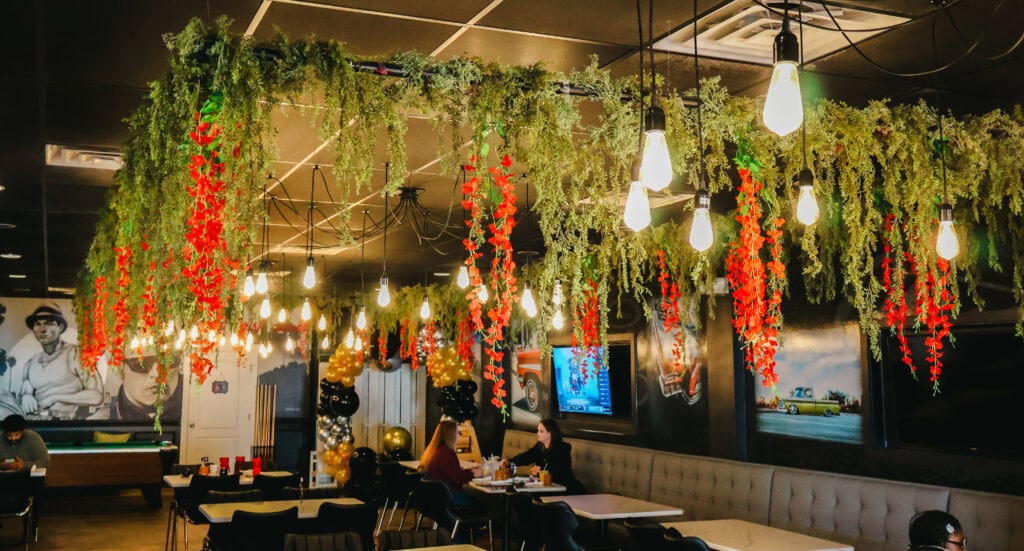 Cozy interior of Chicano's Street Kitchen with lush hanging greenery, red flowers, pendant lights, and diners seated beneath a warm, inviting canopy.