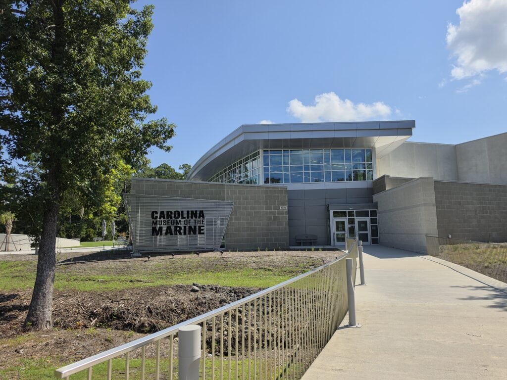 The modern entrance to the Carolina Museum of the Marine in Jacksonville, NC sits beneath a sweeping silver roof and a broad wall of reflective windows on a bright sunny day. Large black lettering on a metal sign beside the walkway reads "CAROLINA MUSEUM OF THE MARINE".