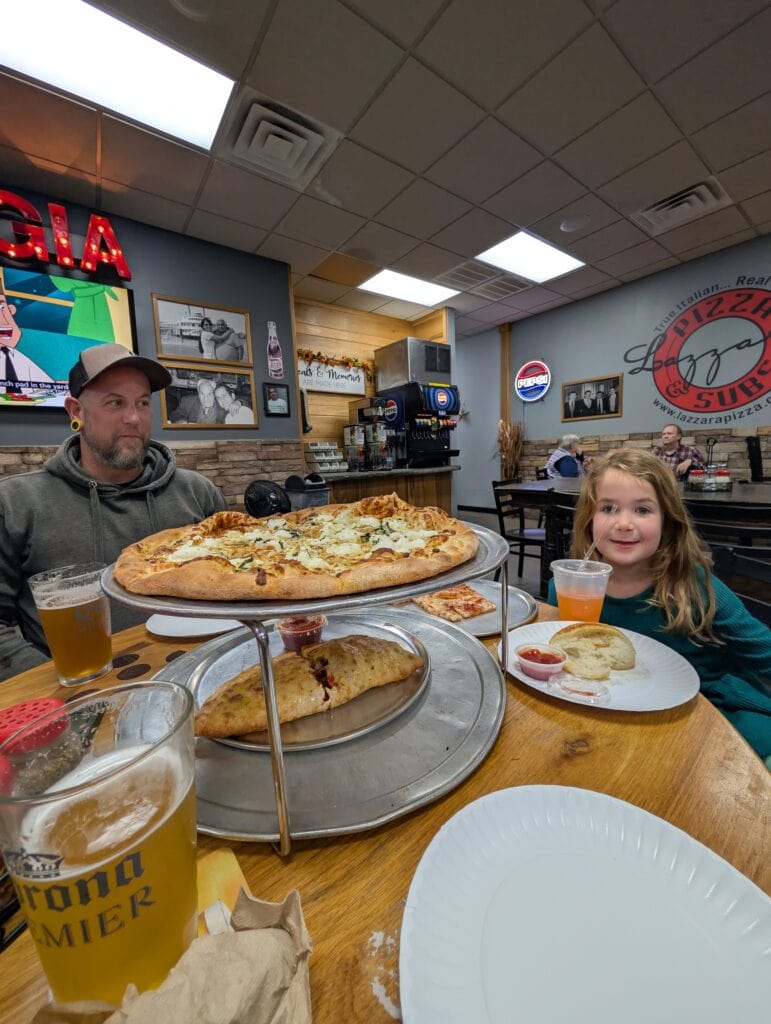 A family enjoys pizza together at Lazzaras Pizza and Subs in Jacksonville NC. A large cheese pizza rests on a raised tray beside a golden baked calzone while a man and smiling young girl sit across the table in the casual Lazzara's Pizza and Subs in Jacksonville, NC.
