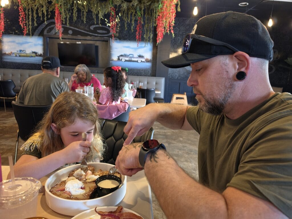 A father and his young daughter sit together at a table at Chicano's Street Kitchen in Jacksonville, NC sharing a large sweet breakfast dish topped with whipped cream fruit and syrup. The girl concentrates on cutting into the food while the man points toward the plate and other diners sit beneath hanging greenery and warm lights in the background.