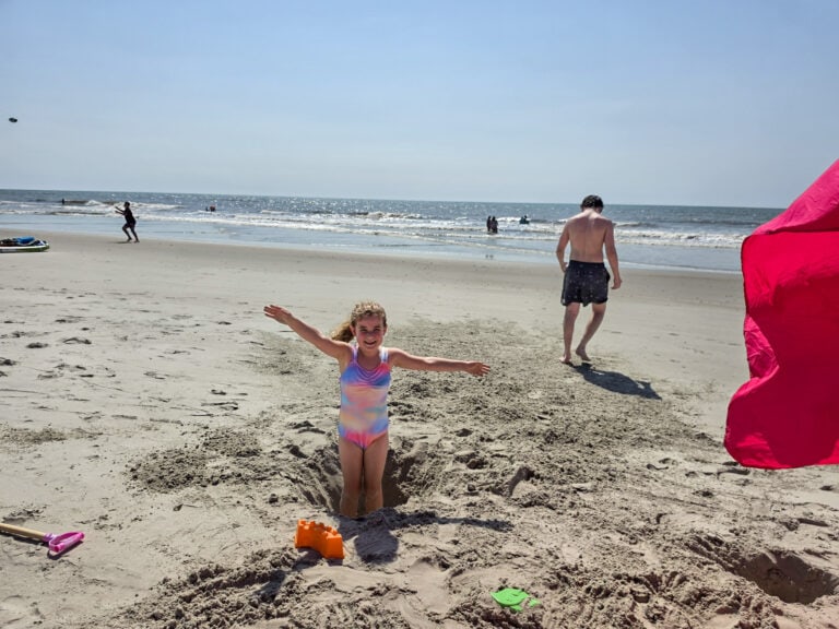 A young girl in a pastel rainbow swimsuit stands smiling with her arms stretched wide inside a deep hole dug in the sand on Bear Island in North Carolina. Her brother walks toward the sparkling ocean behind her while beach toys lie scattered across the sand and swimmers play in the waves at the beach on Bear Island in North Carolina. Featured image for the article Fun, Sun, and History: 2 Days in Jacksonville NC with Kids.