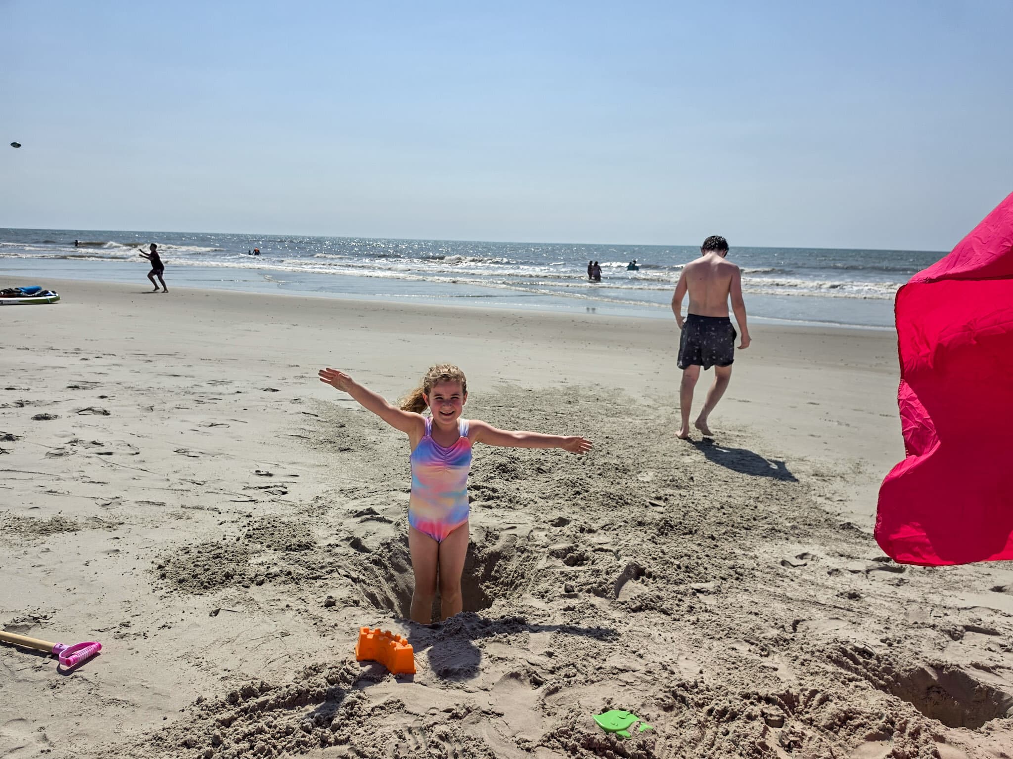 A young girl in a pastel rainbow swimsuit stands smiling with her arms stretched wide inside a deep hole dug in the sand on Bear Island in North Carolina. Her brother walks toward the sparkling ocean behind her while beach toys lie scattered across the sand and swimmers play in the waves at the beach on Bear Island in North Carolina. Featured image for the article Fun, Sun, and History: 2 Days in Jacksonville NC with Kids.