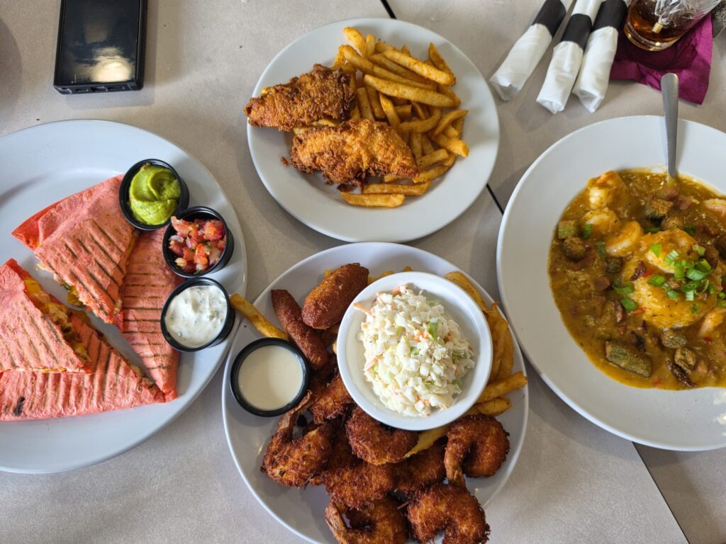 Lunch at Prestons Corner Bar and Grill in Swansboro, North Carolina fills the table with colorful comfort food including grilled wrap wedges with guacamole and pico de gallo. Other plates hold crispy fried seafood with fries and coleslaw. Fried chicken tenders with seasoned fries. And a rich shrimp and okra dish topped with green onions.