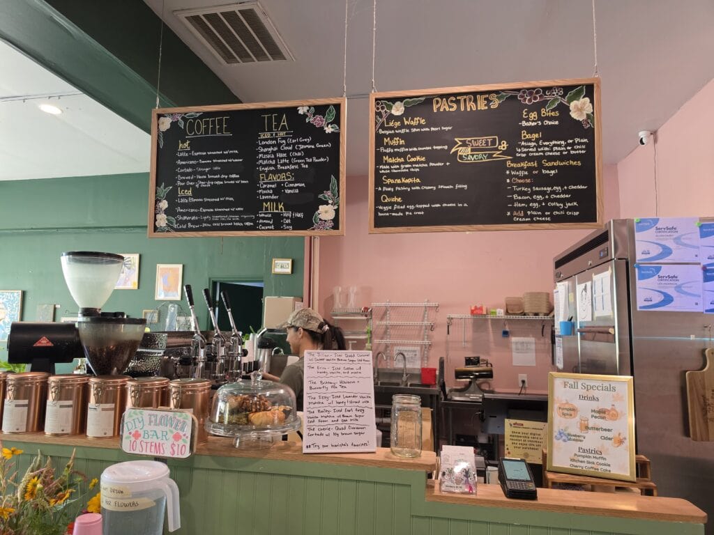 A cozy cafe counter at Milk Road Downtown in Jacksonville, NC with sage green paneling sits beneath large hand drawn chalkboard menus against green and blush pink walls. The boards read "COFFEE" "TEA" and "PASTRIES" while smaller signs advertise "D.I.Y. FLOWER BAR 10 STEMS $10" and "Fall Specials" including pumpkin spice maple pecan butterbeer blueberry cobbler and cider.
