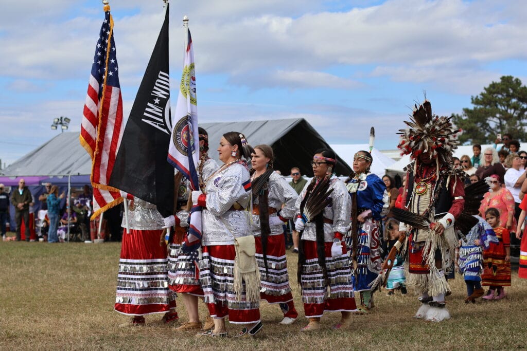 A procession of adults and children in elaborate feathered beaded and brightly patterned regalia at the Onslow Veterans Pow Wow in Jacksonville, NC walks across a grassy festival field while spectators watch from behind. At the front of the group are the United States flag a black "POW MIA" flag and another ceremonial flag.