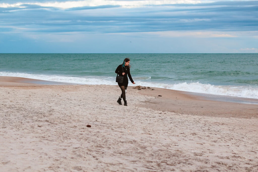 Phoenyx walks along the sandy shoreline at North Topsail Beach with ocean waves behind her. She wears a black outfit and carries a handbag while her prosthetic leg is visible as she explores the beach during a 2 day itinerary in Jacksonville NC.