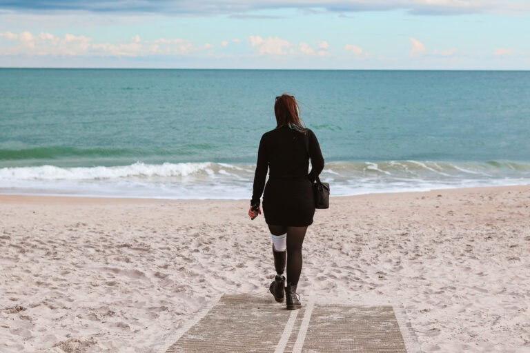 Phoenyx walks toward the shoreline on an accessible beach mat at North Topsail Beach in North Carolina during her 2 day itinerary in Jacksonville, NC. She carries a black handbag and walks with a prosthetic leg across the sandy beach toward the calm blue ocean.