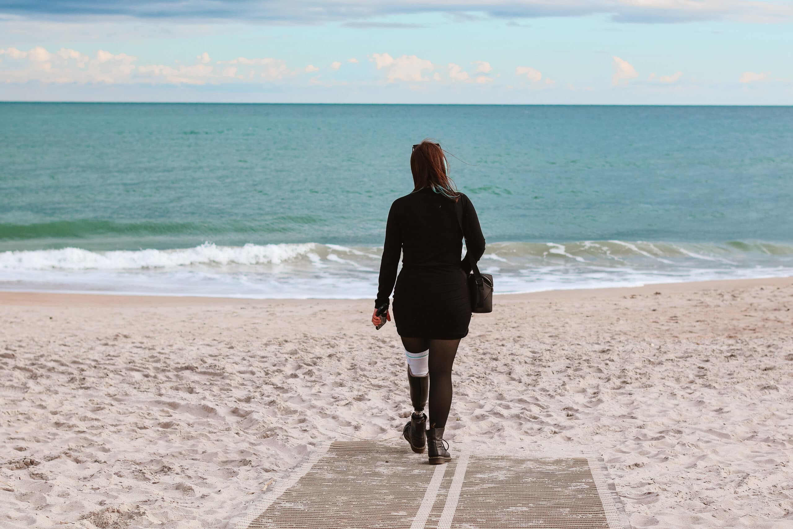 Phoenyx walks toward the shoreline on an accessible beach mat at North Topsail Beach in North Carolina during her 2 day itinerary in Jacksonville, NC. She carries a black handbag and walks with a prosthetic leg across the sandy beach toward the calm blue ocean.