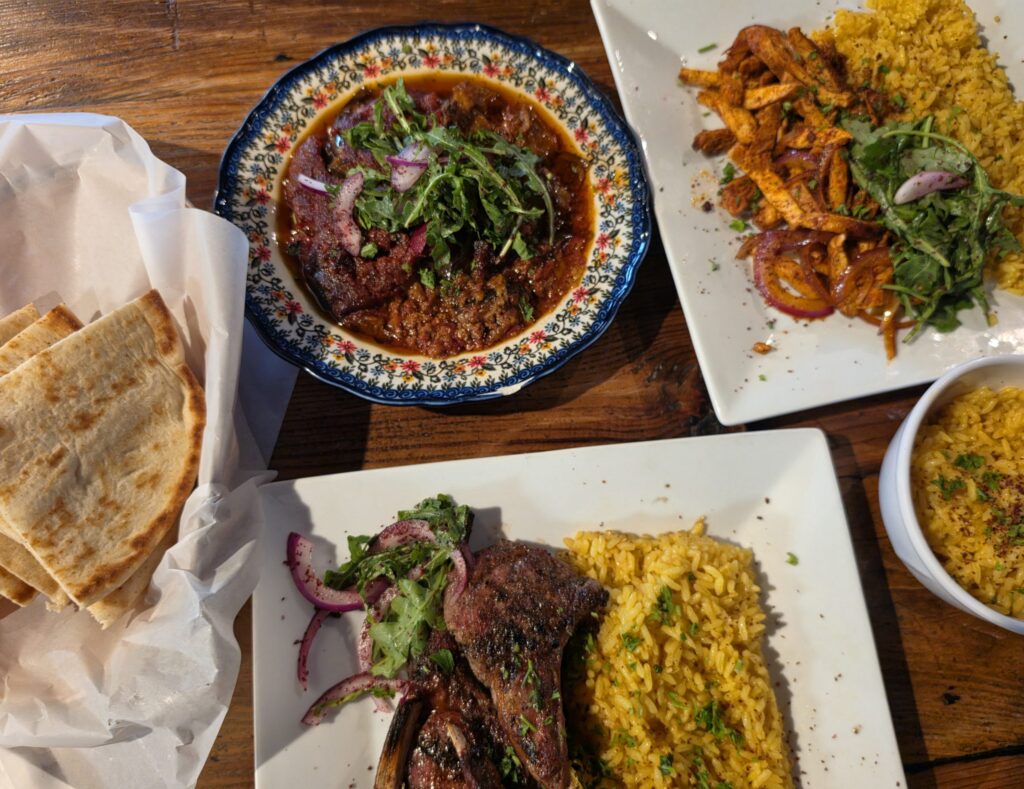 A table is covered with Mediterranean style dishes from Marrakesh restaurant in Jacksonville, NC including grilled meat over yellow rice and seasoned chicken with salad. A patterned bowl holds a saucy meat dish topped with greens and a basket of warm flatbread sits nearby.