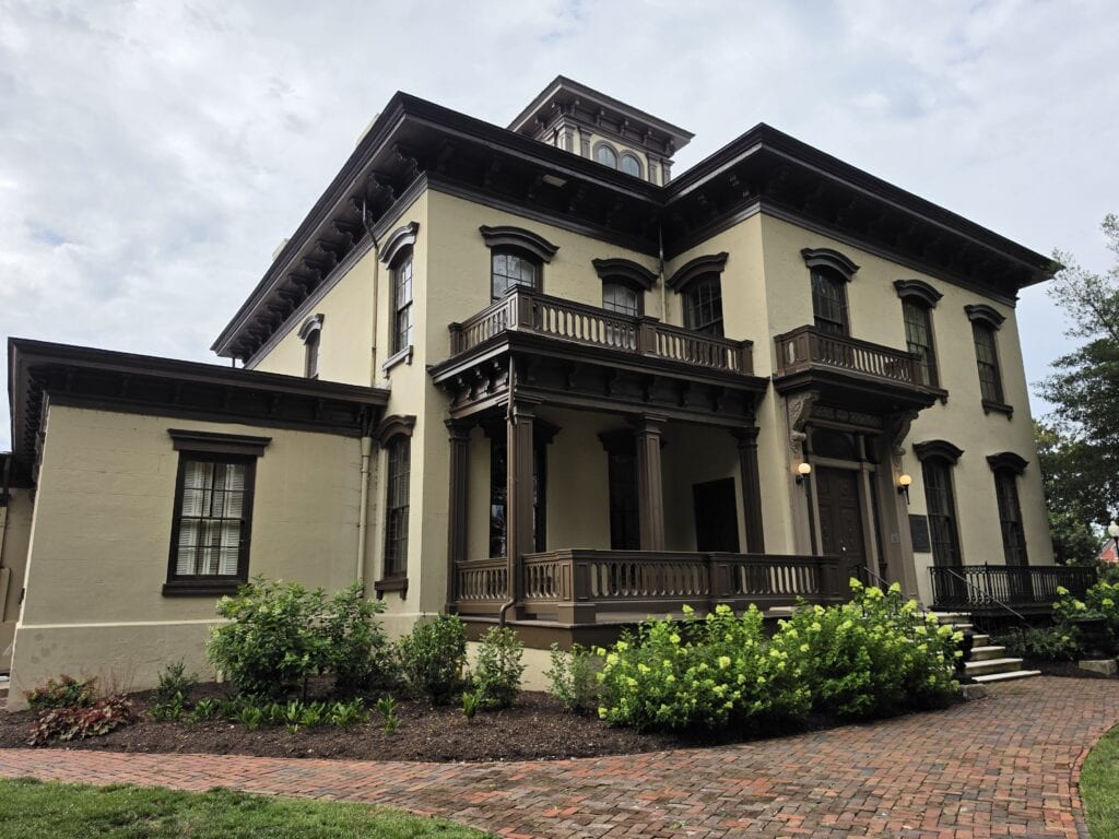 The Danville Museum of Fine Arts and History in Danville VA is housed in a large historic cream colored mansion with dark brown trim. Tall windows decorative balconies and a covered front porch overlook brick walkways and landscaped gardens making the museum one of the historic things to do in Danville VA.